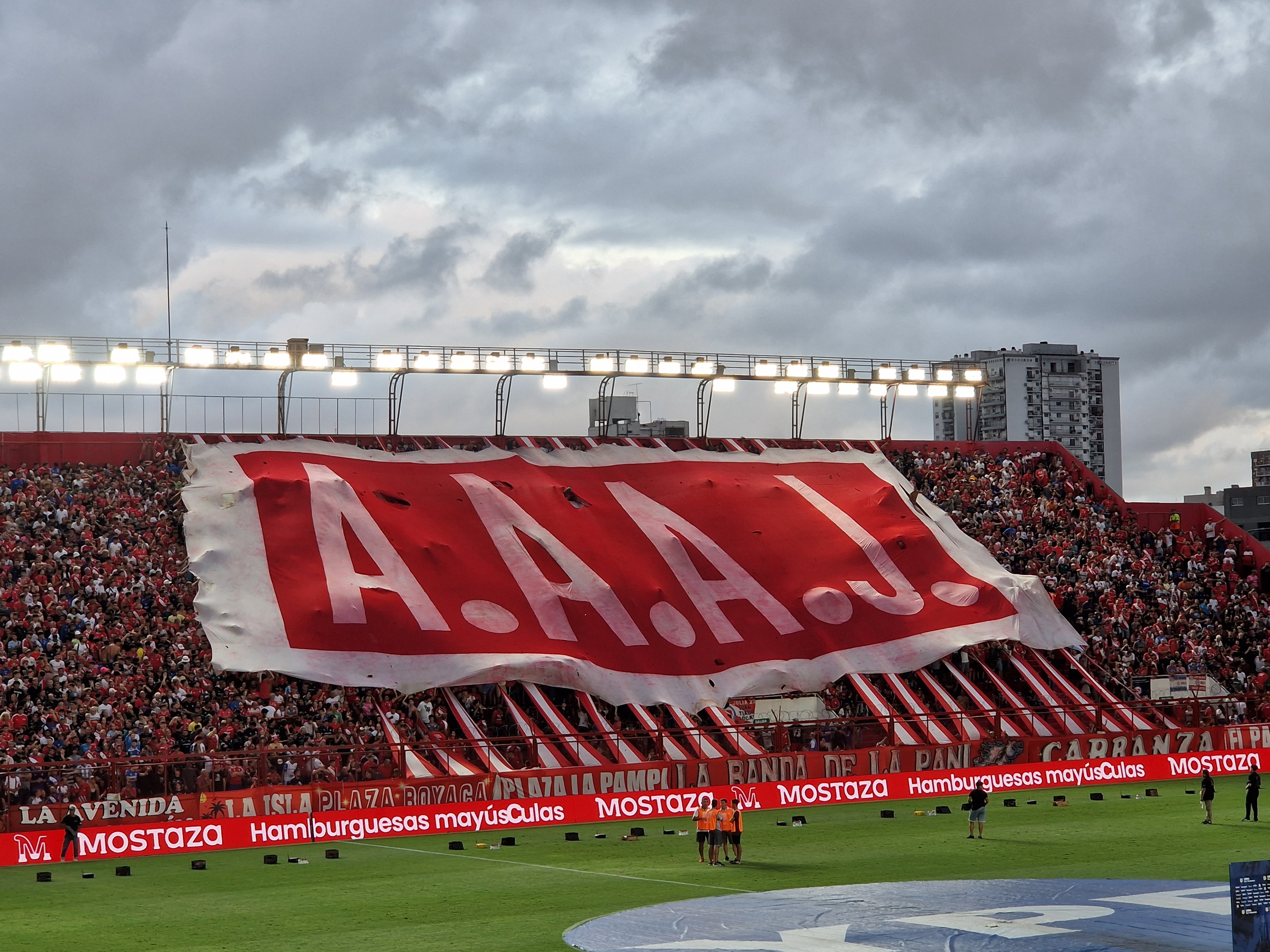 Large red and white banner with 'A.A.A.J.' in the Diego Armando Maradona stadium during an Argentinos Juniors match.
