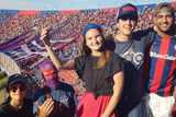 A group of football fans at a match, with one person in the foreground making a peace sign. The fans are dressed in team jerseys and hats, and the atmosphere appears lively and festive.