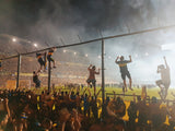 Boca Juniors fans climb the alambrado fence pitchside at a night game in the Bombonera with smoke in the background